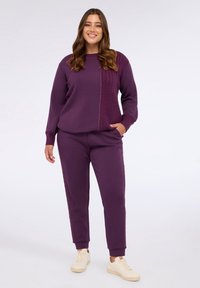 Violetter Sweatshirt- und Jogginganzug. Das Oberteil hat ein strukturiertes Panel und eine lockere Passform; die Hose hat elastische Bündchen und seitliche Taschen.