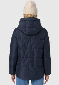 Marineblaue Steppjacke mit Kapuze, mit gestepptem Textur, leichtem Glanz und einem Logo-Patch über dem Saum. Das Model trägt eine beige Strickmütze.