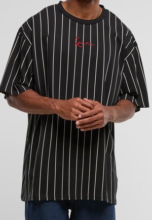 Homme portant un T-shirt noir à fines rayures blanches verticales et un logo rouge en écriture cursive sur la poitrine, les mains jointes devant.