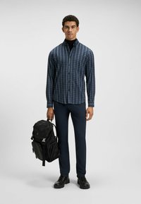 Marineblauw en grijs geruite button-up shirt, slim-fit donkerblauwe broek, zwarte rugzak en zwarte schoenen. Model staat, houdt de rugzak vast.