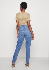 Mujer vestida con una blusa de rayas de manga corta, jeans de tiro alto en color azul y sandalias de tacón oliva, de pie frente a un fondo simple.