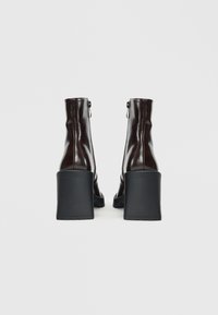 Braune Lackleder-Stiefeletten mit einem klobigen schwarzen Blockabsatz, die eine glatte Textur und einen Reißverschluss an der Seite aufweisen.