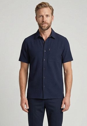 Man met kort blond haar en baard, gekleed in een donkerblauw overhemd met korte mouwen en bijpassende broek, tegen een effen achtergrond.