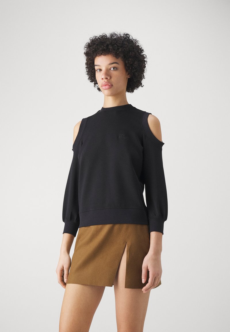 Sudadera negra de hombros al descubierto con mangas largas y abullonadas, y dobladillo acanalado, combinada con una falda mini marrón con una abertura frontal.