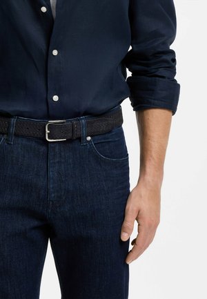 Mężczyzna ubrany w ciemnoniebieską koszulę z guzikami, z podwiniętymi rękawami, ciemnoniebieskie dżinsy oraz czarny pleciony skórzany pasek z srebrną klamrą.