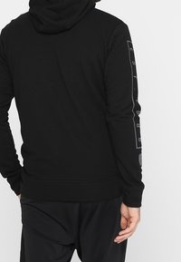 Sudadera negra hecha de una tela suave, con un diseño clásico, capucha ajustable con cordón y detalle impreso a lo largo de la manga.
