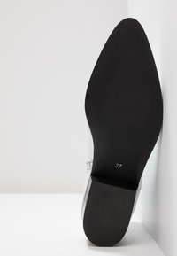 Suela de un zapato blanco con punta afilada, fondo texturizado negro, tacón cuadrado y número de talla 37 visible cerca del arco.
