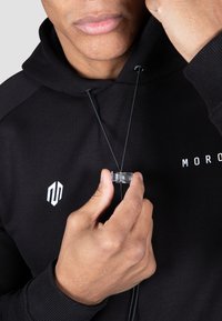 Hanorac negru cu un logo alb, având un șnur de ajustare și un mecanism de blocare transparent, ținut într-o mână. Materialul pare moale, cu o textură fină.