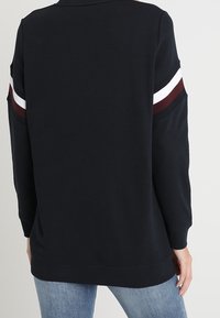 Persona con una camiseta de manga larga negra con rayas blancas y burdeos en las mangas, combinada con unos jeans de mezclilla azul claros, vista desde atrás.