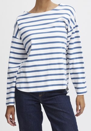 Personne portant une chemise blanche à manches longues avec des rayures horizontales bleues et un jean en denim bleu foncé, debout les mains détendues.