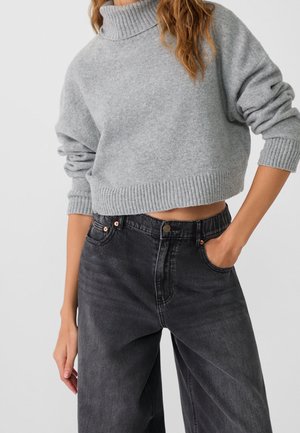 Personne portant un pull court gris à col roulé avec manches longues et un jean noir taille haute, une main dans la poche.