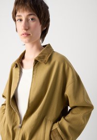 Light olive green zip-up jacket with a corduroy collar, large pockets, dropped shoulders, and a soft textured finish.