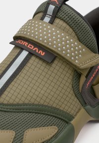 Κοντινή λήψη από sneaker σε ελιά πράσινο και ταμπά με υφή, που διαθέτει ανακλαστική λωρίδα, τρυπητό λουράκι και κόκκινο λογότυπο "Jordan" στο λουράκι και στην γλώσσα.