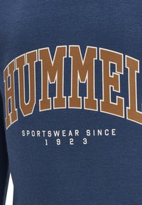 Blaues Sweatshirt mit auffälliger goldener Beschriftung "HUMMEL" und "SPORTSWEAR SINCE 1923" in Weiß darunter. Weicher, strukturierter Stoff.