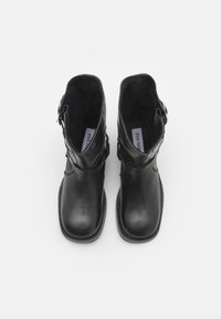 Botas de tobillo de cuero negro con un acabado suave, que presentan un detalle de hebilla en el lateral y una puntera redondeada. Forro interior suave visible.