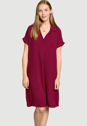 Jeune femme aux longs cheveux blonds porte une robe bordeaux ample à manches courtes avec un col et un décolleté en V, sur un fond clair.