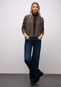 Bruine gewatteerde bomberjack met een zwarte coltrui, gecombineerd met blauwe jeans met wijde pijpen en zwarte laarzen. Model staat met handen in de zakken.