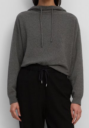 Personne portant un sweat à capuche en maille grise avec cordons de serrage et un pantalon noir à taille élastique avec cordon, debout devant un fond clair uni.