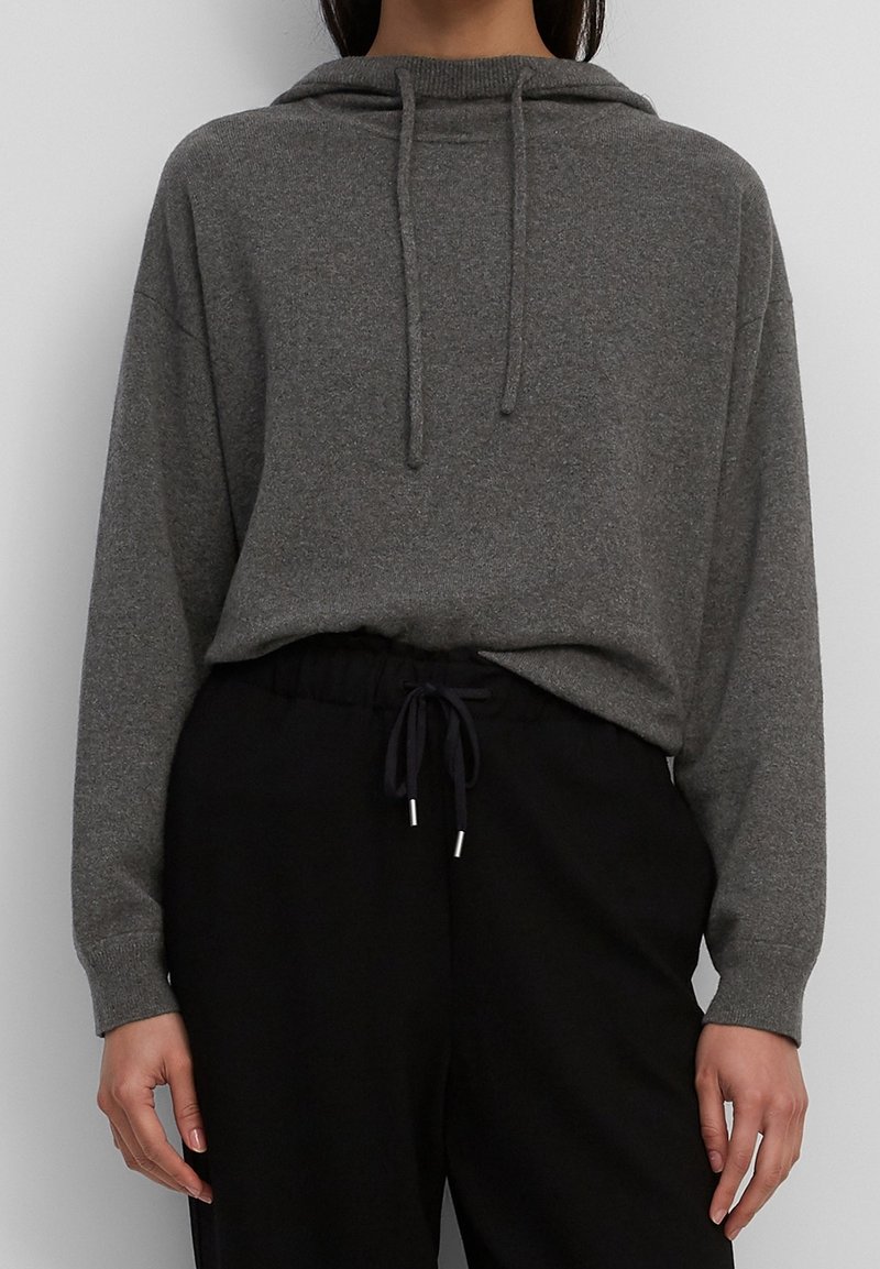 Personne portant un sweat à capuche en maille grise avec cordons de serrage et un pantalon noir à taille élastique avec cordon, debout devant un fond clair uni.