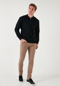 Fekete zipzáras kapucnis pulóver hosszú ujjakkal, bézs slim-fit nadrág, és fekete tornacipő fehér talppal. Simaságú textúra, laza illeszkedés.