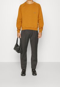Sudadera color mostaza con mangas raglán, pantalones oscuros de sastre, bolso de mano acolchado negro y zapatos de cuero negros.