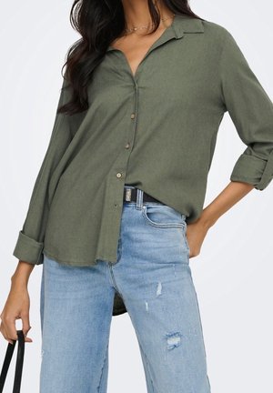 Femme portant une chemise boutonnée vert olive avec manches retroussées, rentrée dans un jean bleu clair déchiré, tenant la bandoulière d’un sac à main noir.