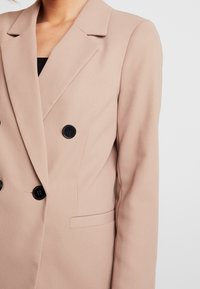 Blazer beige de doble botonadura con botones negros, con cuello de solapa, textura suave y bolsillo lateral. Corte entallado con silueta estructurada.