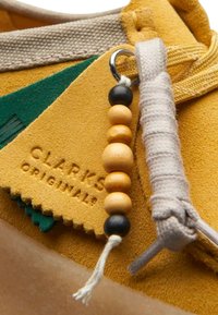 Gul mockaskor med gröna detaljer, med en texturerad etikett präglad med "CLARKS ORIGINALS," och dekorerade pärlor i svart och träfärger.