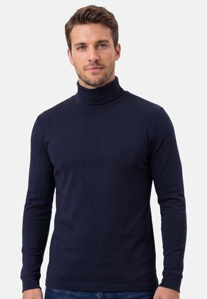 Mann mit kurzen braunen Haaren, der einen taillierten marineblauen Rollkragenpullover und Jeans trägt und vor einem schlichten hellen Hintergrund steht.
