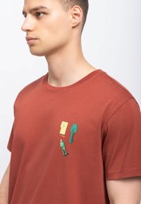 Maglietta rossa in cotone con scollatura rotonda che presenta tre applicazioni grafiche colorate: giallo, verde e una piccola figura, aggiungendo un dettaglio visivo.