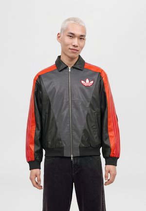 Un mannequin aux cheveux courts décolorés porte une veste noire zippée avec des manches à rayures rouge vif et un logo trèfle blanc, sur un fond uni.
