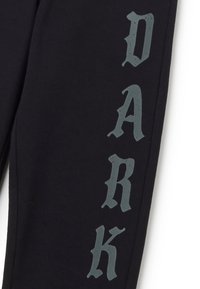 Zwarte joggingsbroek met een gladde textuur heeft grote, versleten grijze letters die "DARK" verticaal langs één been spellen.