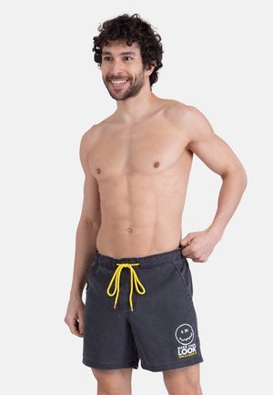 Homme souriant torse nu aux cheveux bouclés, portant un short de bain noir avec un cordon jaune et un graphique visage souriant blanc sur la jambe gauche.