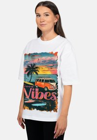 Wit katoenen T-shirt met een kleurrijke afbeelding van een vintage oranje camperbus en palmbomen tegen een zonsondergangachtergrond.