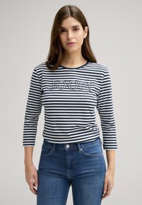 Camicia a righe maniche lunghe in blu navy e bianco con scollatura tonda, con logo "JOOP!" sul davanti. Abbinata a jeans in denim blu.