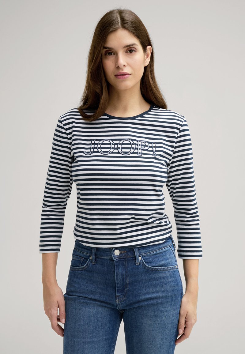 Camicia a righe maniche lunghe in blu navy e bianco con scollatura tonda, con logo "JOOP!" sul davanti. Abbinata a jeans in denim blu.