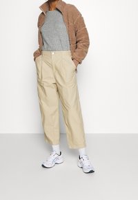 Beige Utility-Hosen mit weitem Bein und frontalen Taschen, kombiniert mit einem grauen Pullover und einer braunen Fleecejacke. Weiße Sportsneaker runden den Look ab.