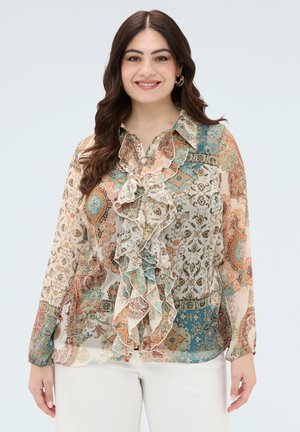 Frau mit langen dunklen Haaren, die eine mehrfarbige florale Paisley-Bluse mit Rüschen und weiße Hosen trägt, lächelnd vor hellem Hintergrund.