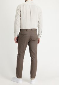 Man wearing beige long-sleeve shirt, brown slim-fit trousers, and white sneakers, standing with back facing the camera on plain background.