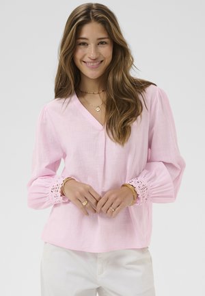 Young woman with long brown hair smiling, wearing a light pink blouse with lace cuffs and white pants, accessorized with gold jewelry.