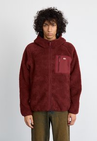 Jachetă din fleece burgundy cu fermoar în față, glugă și buzunar cu clapetă. Material moale, texturat, cu o croială lejeră și manșete elastice.