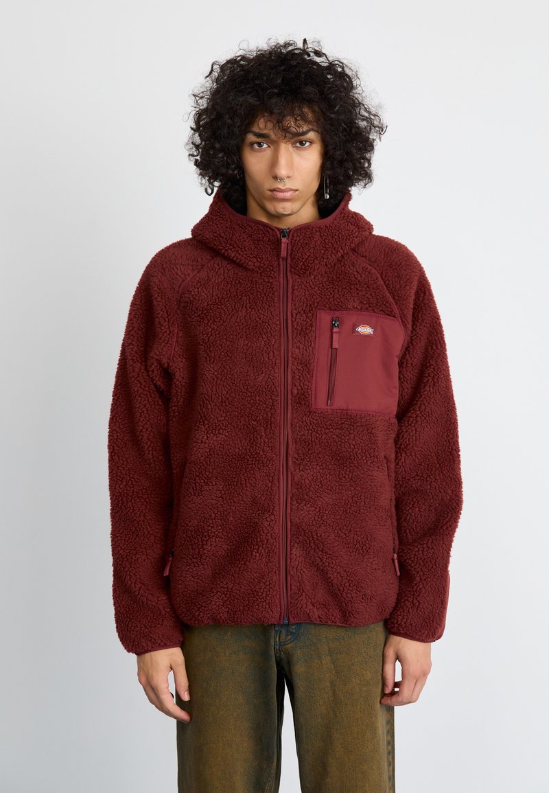 Veste polaire bordeaux avec fermeture éclair à l'avant, capuche et poche plaquée. Matériau doux et texturé avec une coupe décontractée et des poignets élastiques.