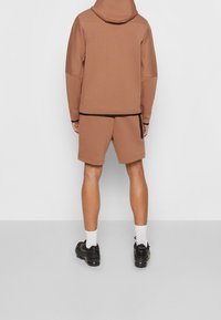 Hnědá mikina s kapucí a odpovídajícími šortkami. Materiál se zdá být měkký s hladkým povrchem. Na lemech a bočních kapsách jsou viditelné černé akcenty.