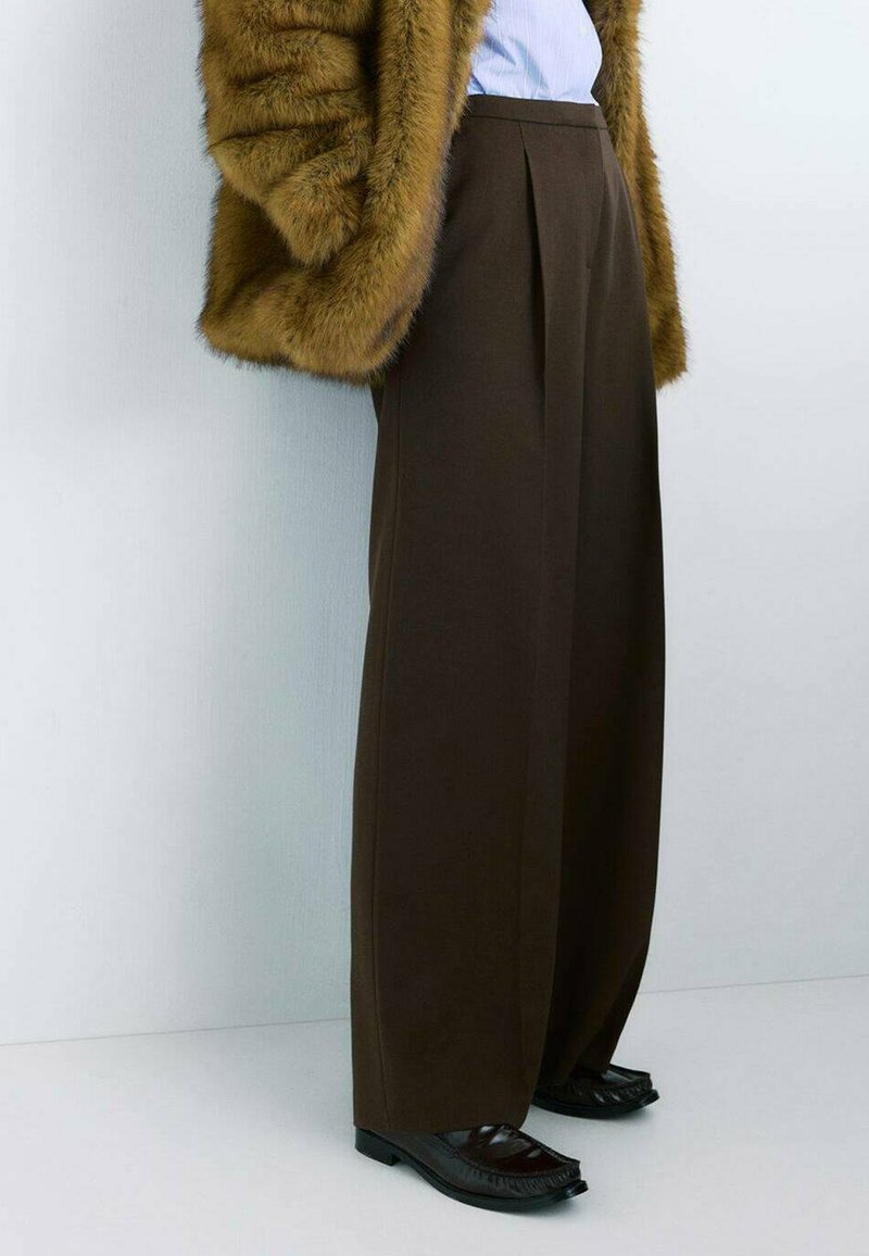 Pantalon plissé marron à coupe ample, en matière douce, associé à des chaussures noires brillantes avec un design texturé.