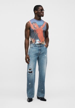 Brezrokavnik z grafičnim potiskom na modrem ozadju, kombiniran s svetlo modrimi obrabljenimi kavbojkami s pravimi hlačnicami in črnimi čevlji.