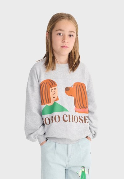 Junge Person mit glattem Haar trägt einen grauen Pullover mit Cartoon-Gesichtern eines Mädchens und eines Hundes sowie eine hellblaue Hose mit kleinem Charakter-Design.