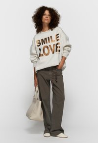 Sudadera gris claro con "SMILE LOVE" en dorado metálico, combinada con pantalones verde oliva y un bolso tote crema. Presenta un ajuste holgado y mangas con puños.