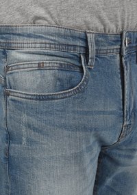 Svijetloplave traperice imaju opušteni kroj s klasičnim dizajnom od pet džepova. Tkanina prikazuje suptilnu, istrošenu teksturu s narančastim šavovima kao akcentima.