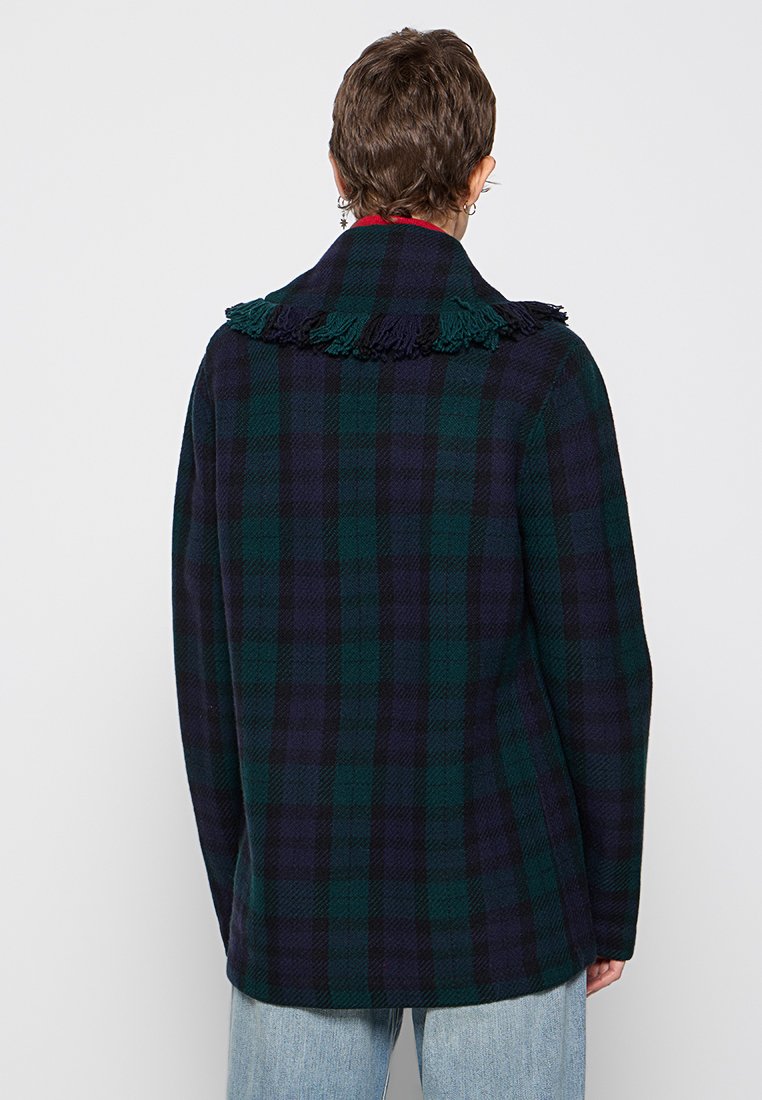 Osoba ubrana w ciemnozieloną i granatową kurtkę w kratę z frędzlami u kołnierza, w połączeniu z jasnoniebieskimi dżinsami, zwrócona plecami w kierunku jednolitego tła.