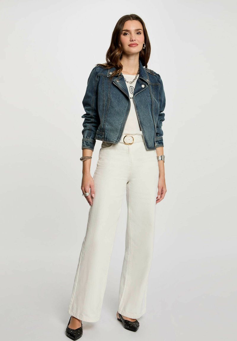 Giacca di jeans con maniche lunghe, colore azzurro chiaro, design aderente. Indossata sopra una T-shirt bianca e abbinata a pantaloni bianchi a gamba larga a vita alta.